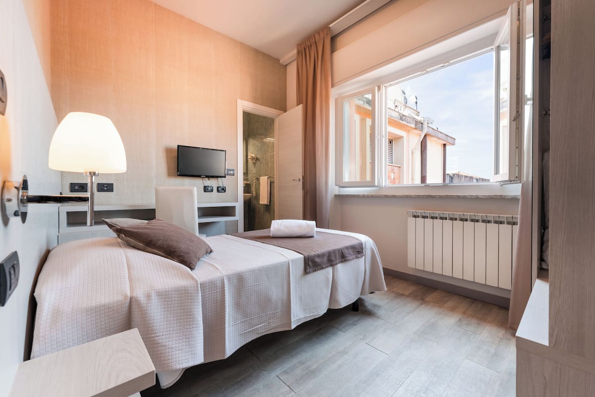 Una habitación de hotel con cama, televisión, ventana y vistas a edificios, además de un radiador.