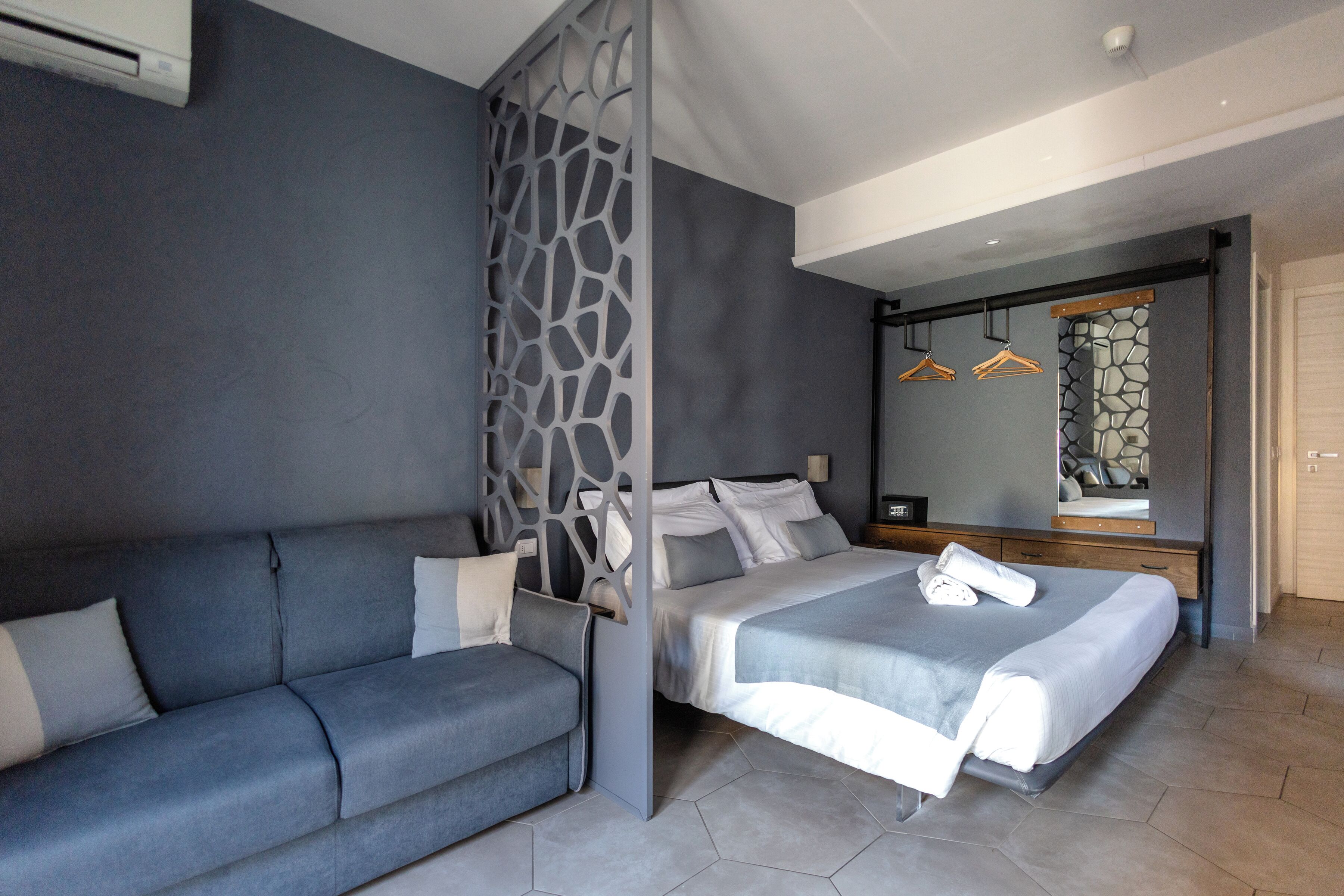 A modern hotel room with a grey sofa, a bed with white linens, a decorative partition, and a mirror with a stone-like backsplash.