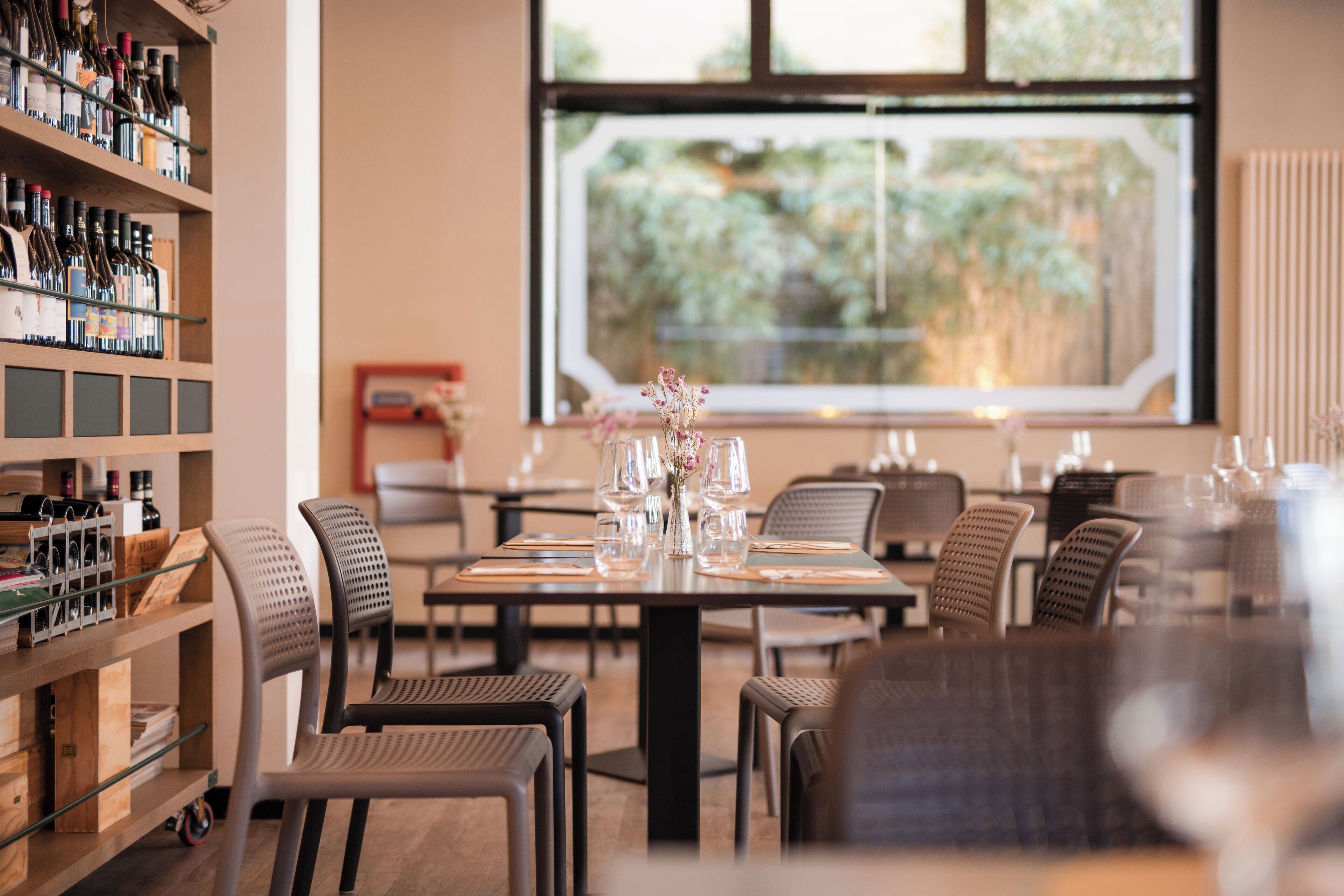 A restaurant interior with a dining table set for four, chairs, a wine rack, and a window with a view of greenery.