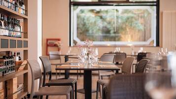 A restaurant interior with a dining table set for four, chairs, a wine rack, and a window with a view of greenery.