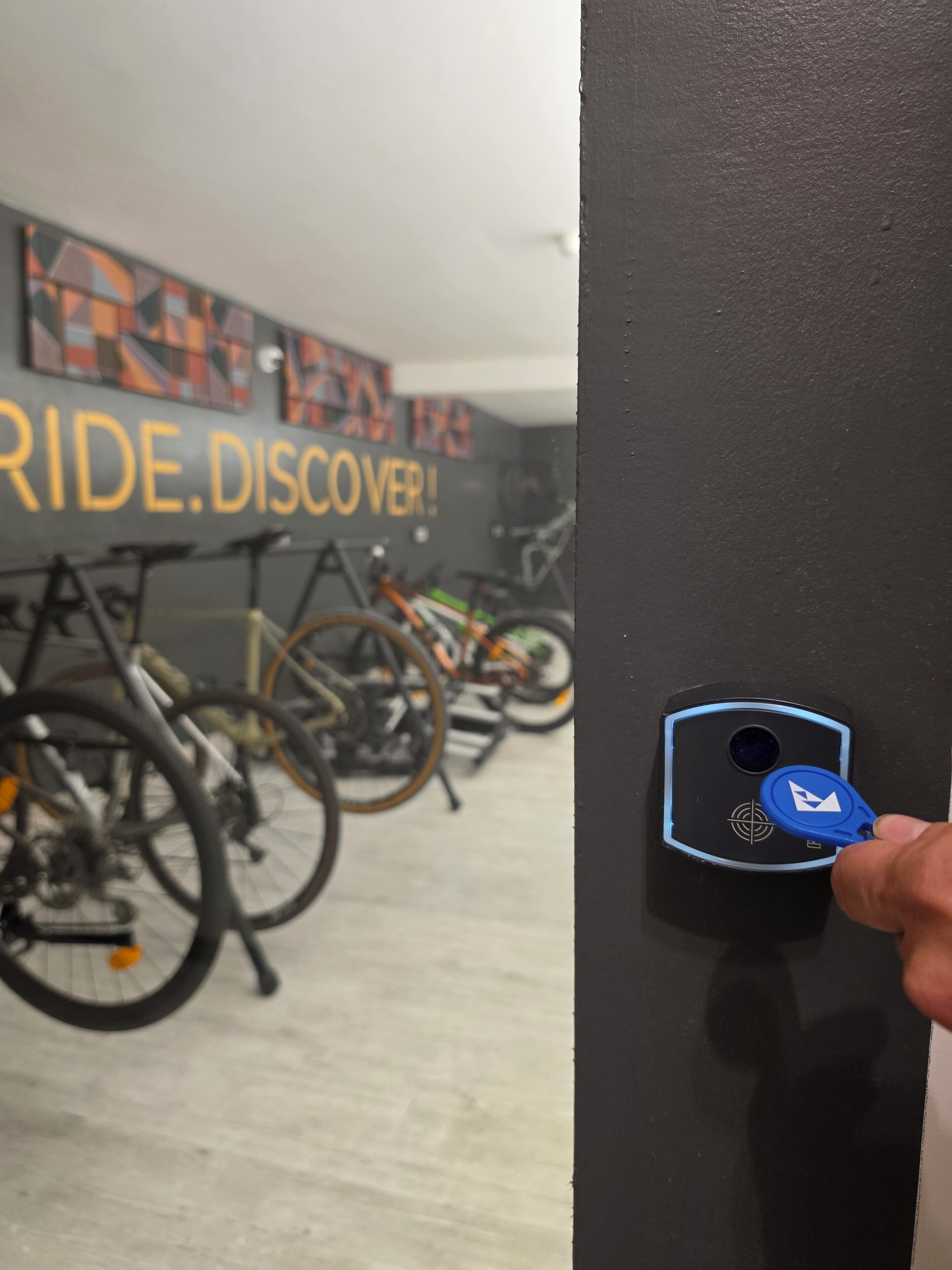 A hand using a keycard to access a door with a sign reading 'RIDE DISCOVER!' in the background, surrounded by bicycles.
