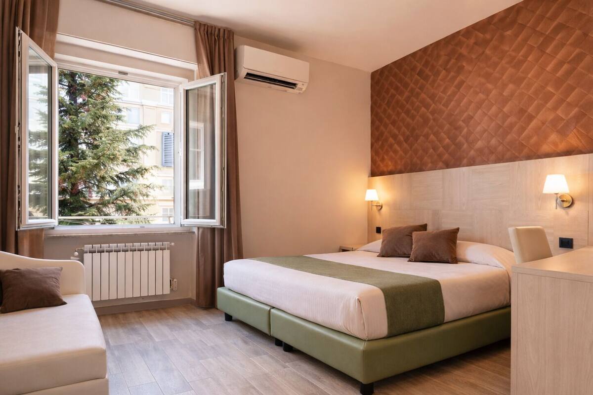 Una habitación de hotel con cama, ventana con cortinas, radiador y lámpara fijada a la pared.