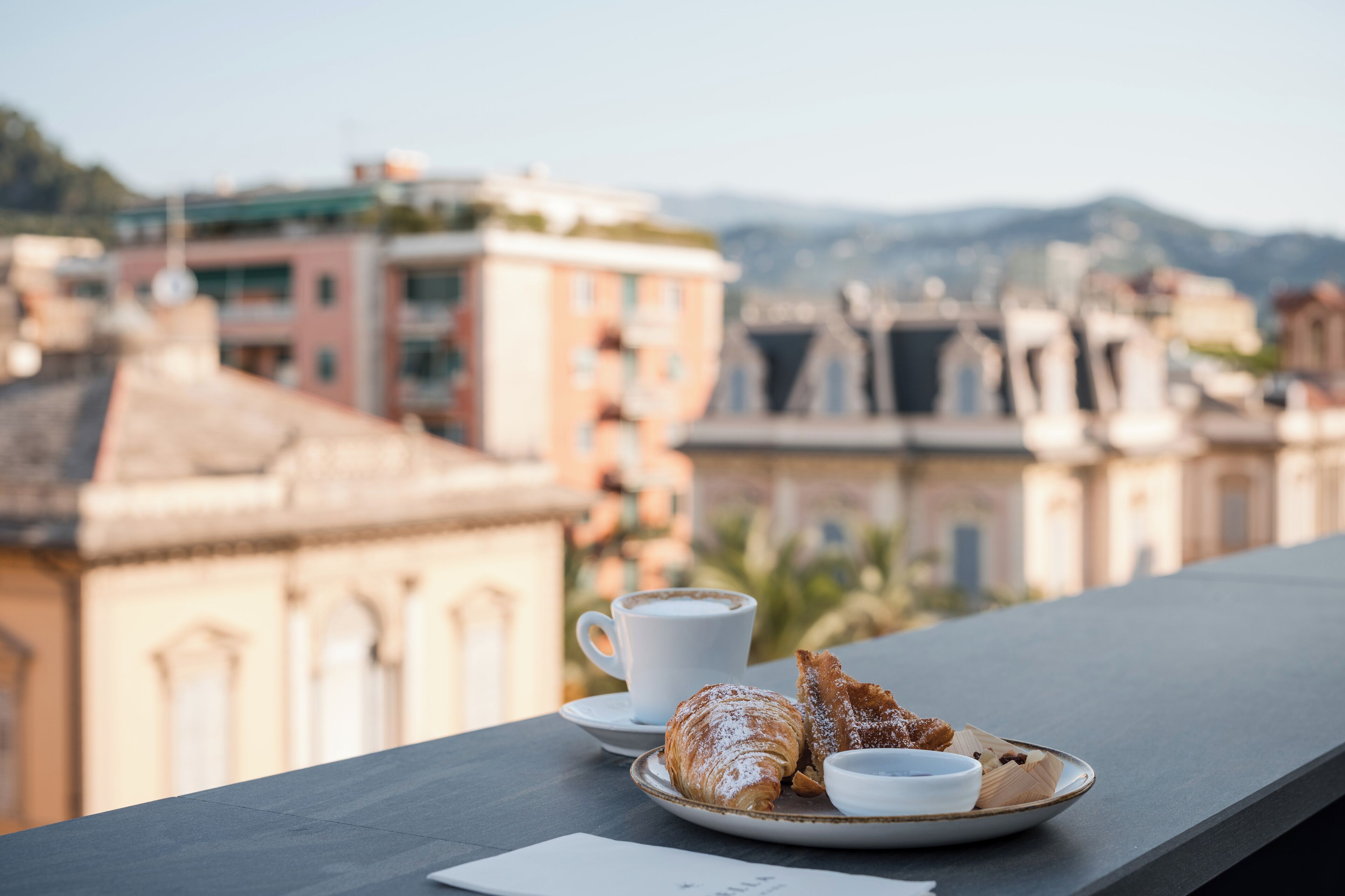 A table with a croissant, a cup of coffee, and a plate with a pastry, overlooking a cityscape with buildings and mountains.