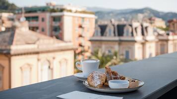 A table with a croissant, a cup of coffee, and a plate with a pastry, overlooking a cityscape with buildings and mountains.