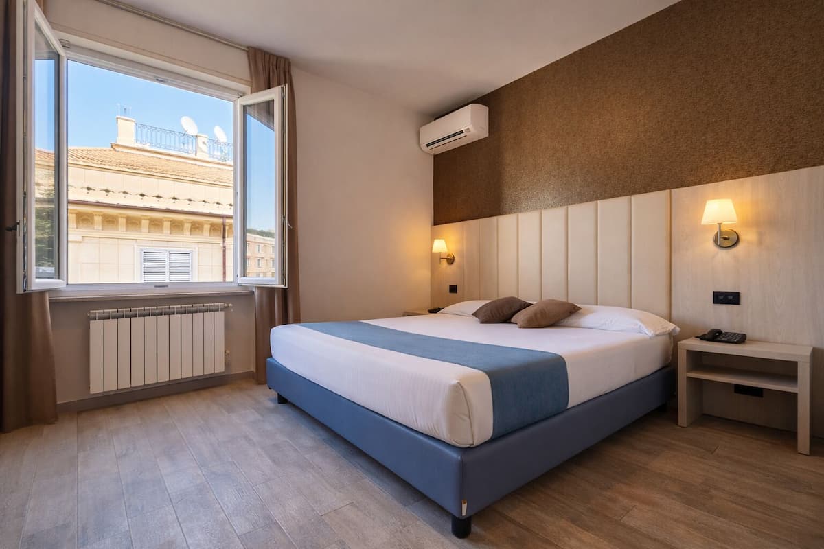 Habitación de hotel con una cama grande, una ventana con cortinas, un radiador, un equipo de aire acondicionado, una mesita de noche con lámpara y una luz en la pared.