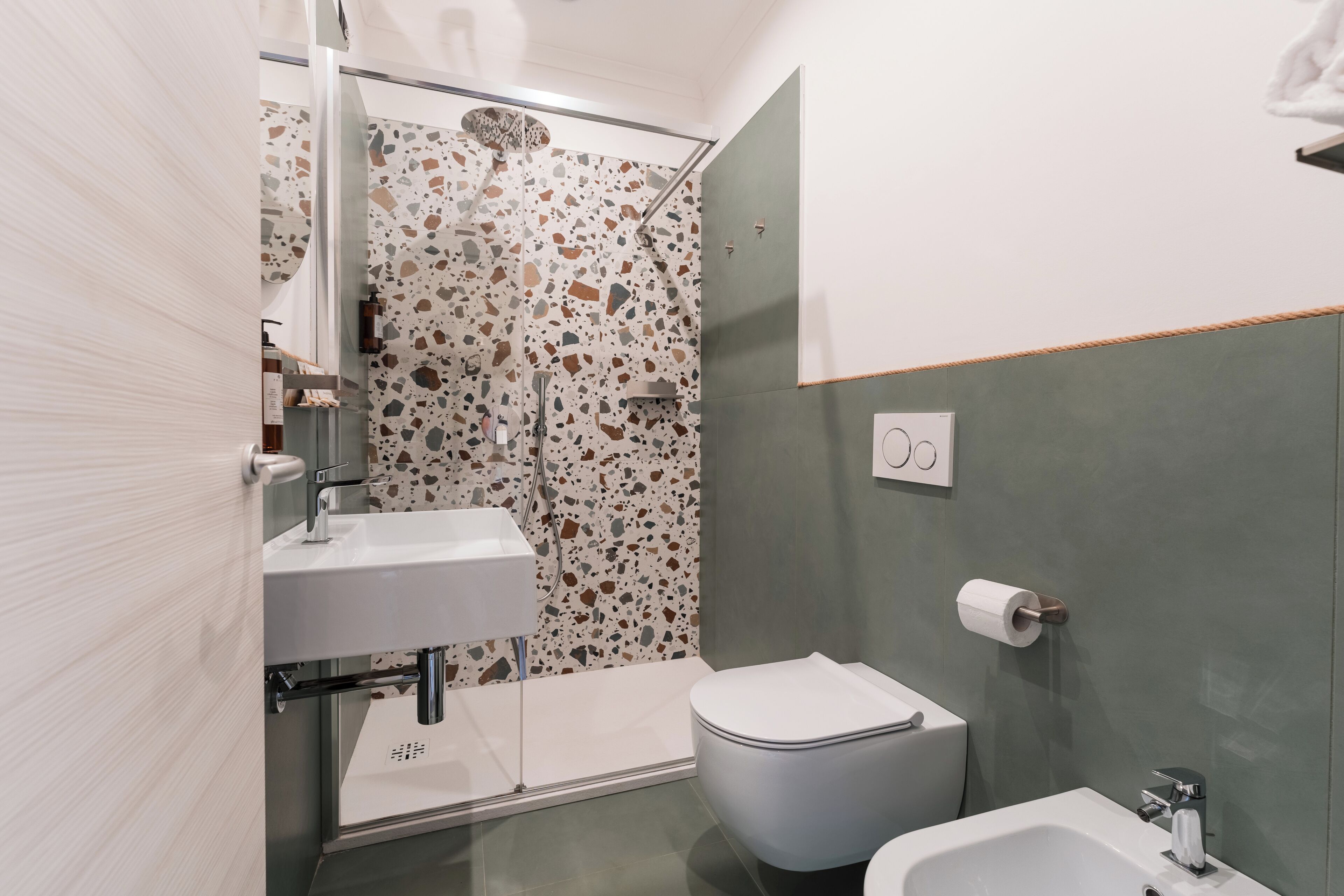 A modern bathroom with a white sink, a toilet, and a bidet. The walls have a terrazzo finish with multicolored stone patterns.