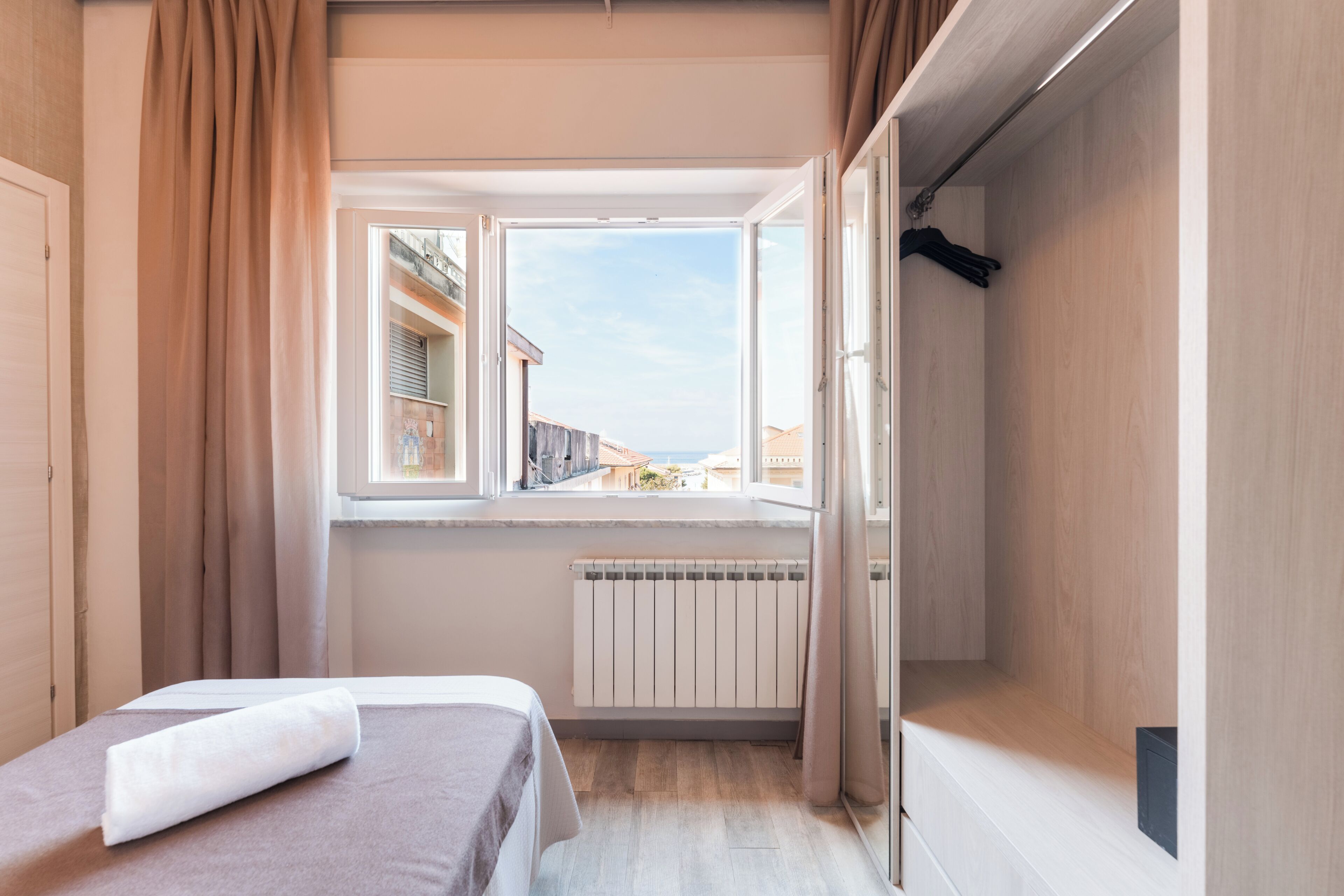 A bedroom with a bed, a window with a view of buildings, a radiator, and a wardrobe.