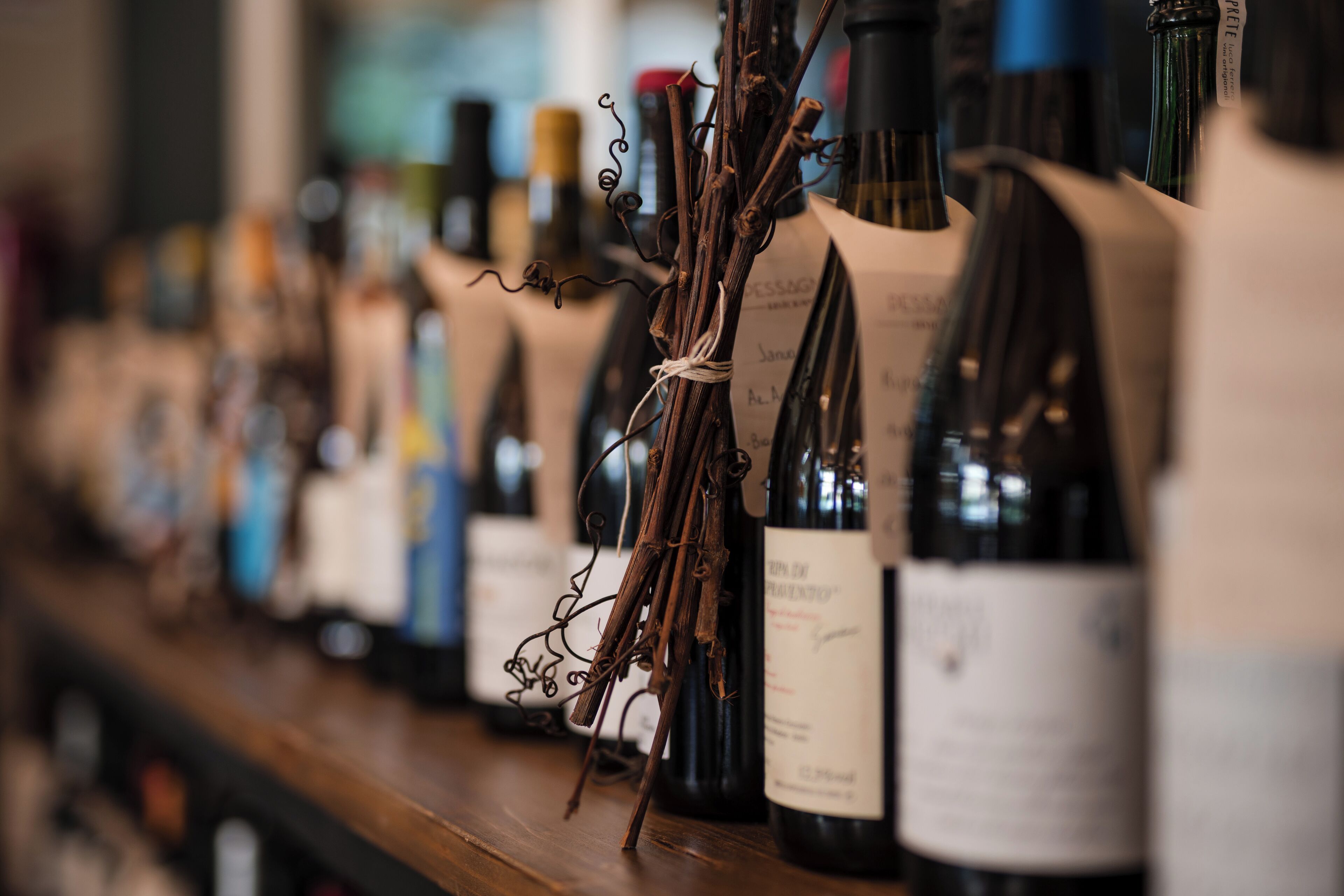 A row of wine bottles on a shelf with labels and twigs tied to them.