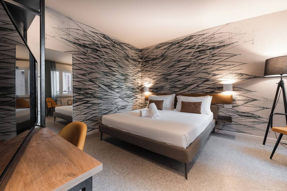 Habitación de hotel moderna con una cama grande, un escritorio y una silla. Hay un espejo en una pared y una lámpara en la otra.