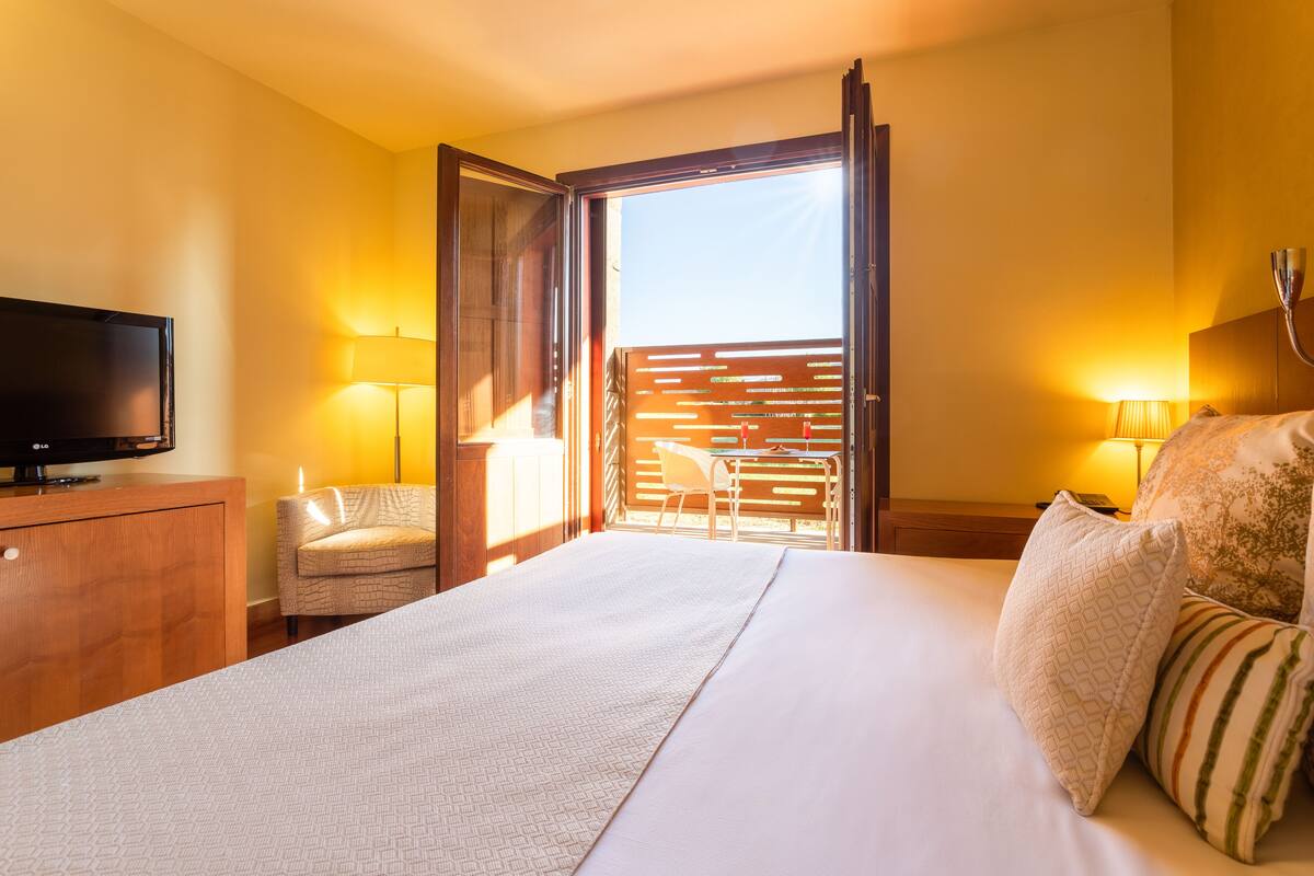 Habitación de hotel con cama, televisión, silla y balcón con vistas.