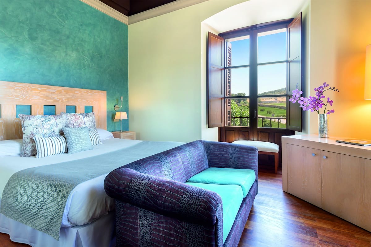 Un dormitorio con cama, sofá, mesita de noche, lámpara, ventana con vistas y un jarrón con flores.