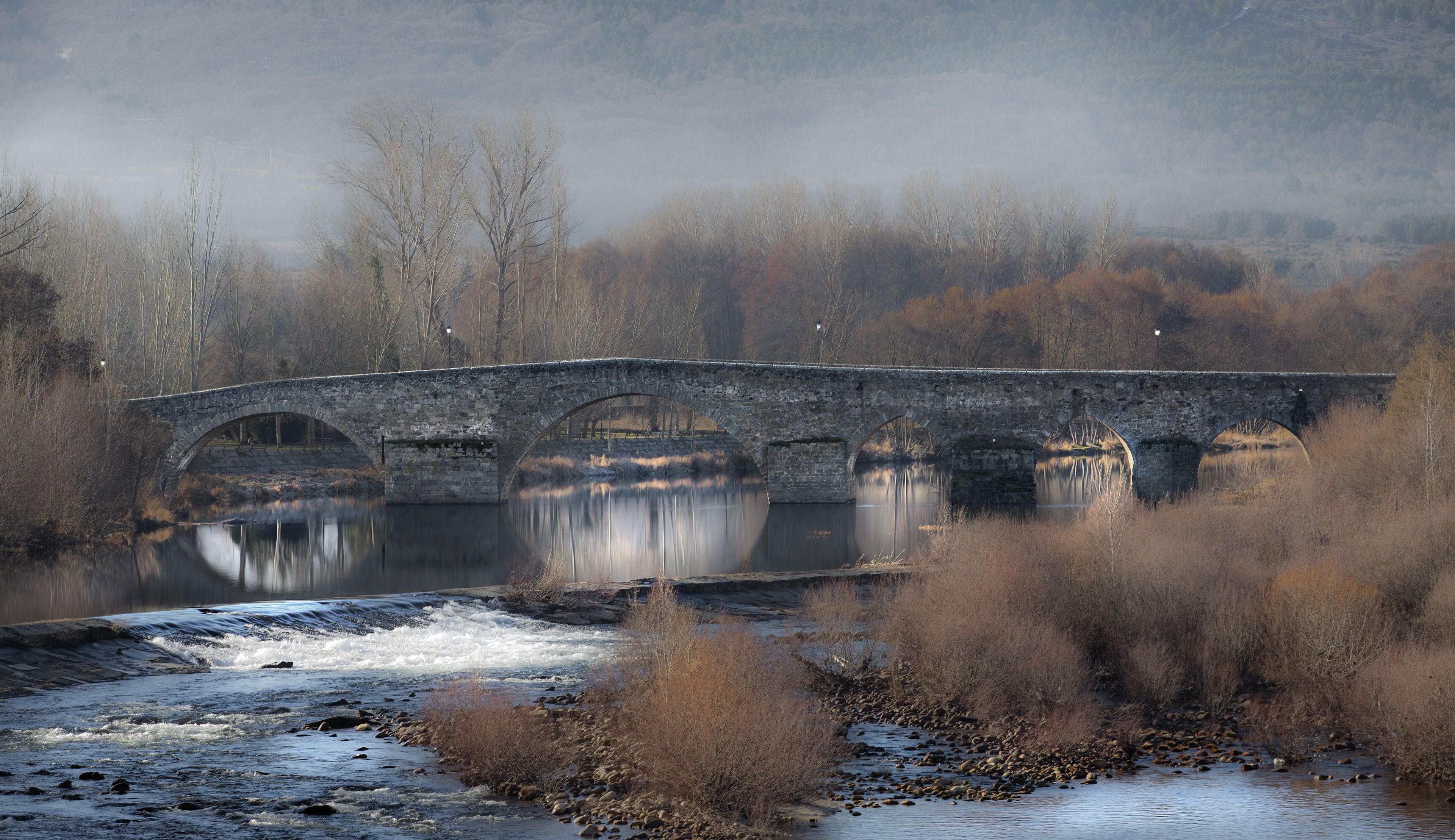 A stone bridge over a calm river with reflections, surrounded by trees and a misty background.