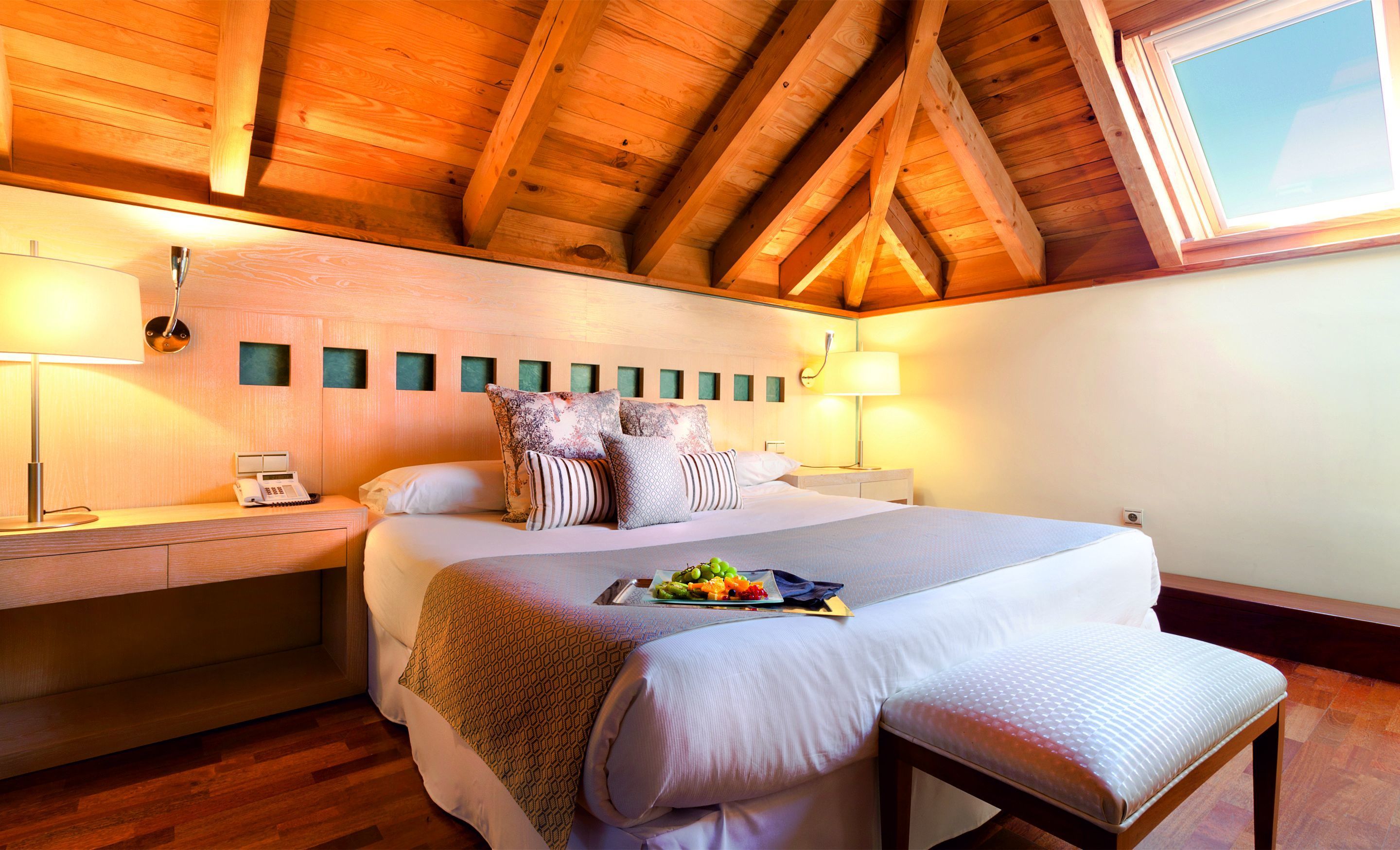 Un dormitorio con techo de madera, una cama con múltiples almohadas, mesitas de noche con lámparas y una ventana.