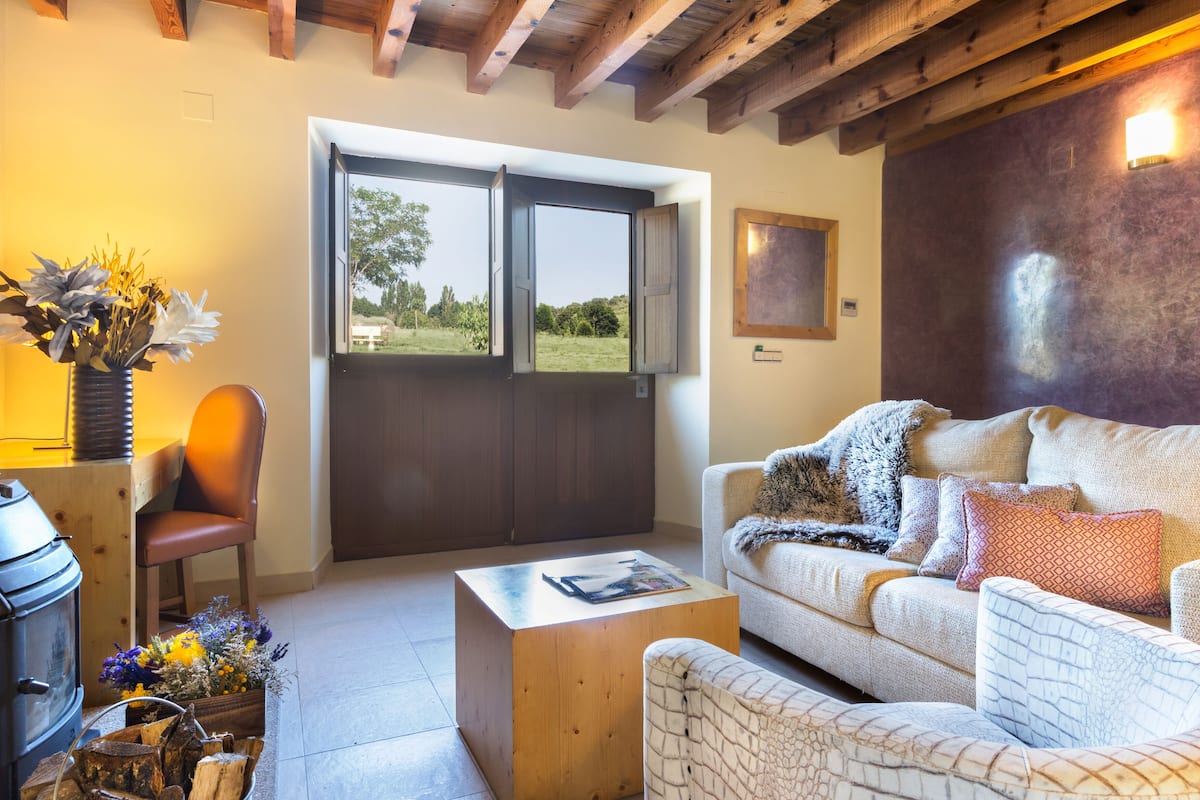 Un salón acogedor con techo de madera, un sofá con cojines estampados, una mesa de centro de madera y vistas a través de un ventanal.