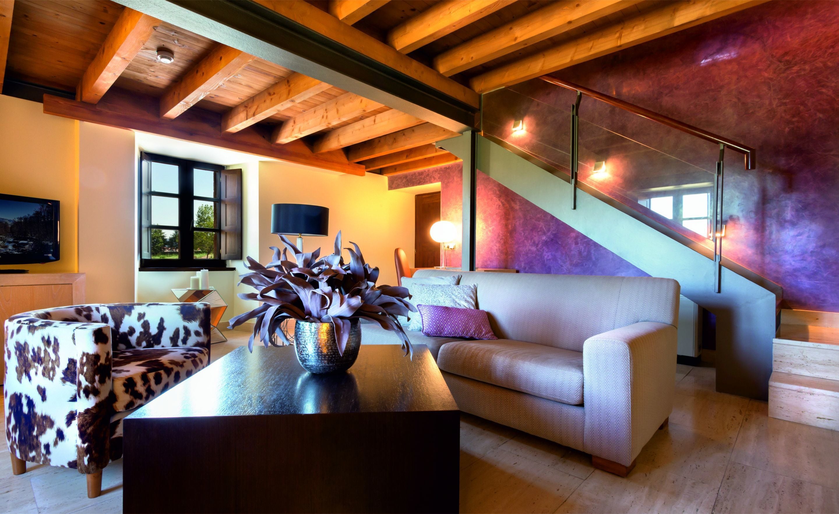 A modern living room with a sofa, a coffee table, and a TV. There is a staircase with a purple wall and a wooden ceiling.