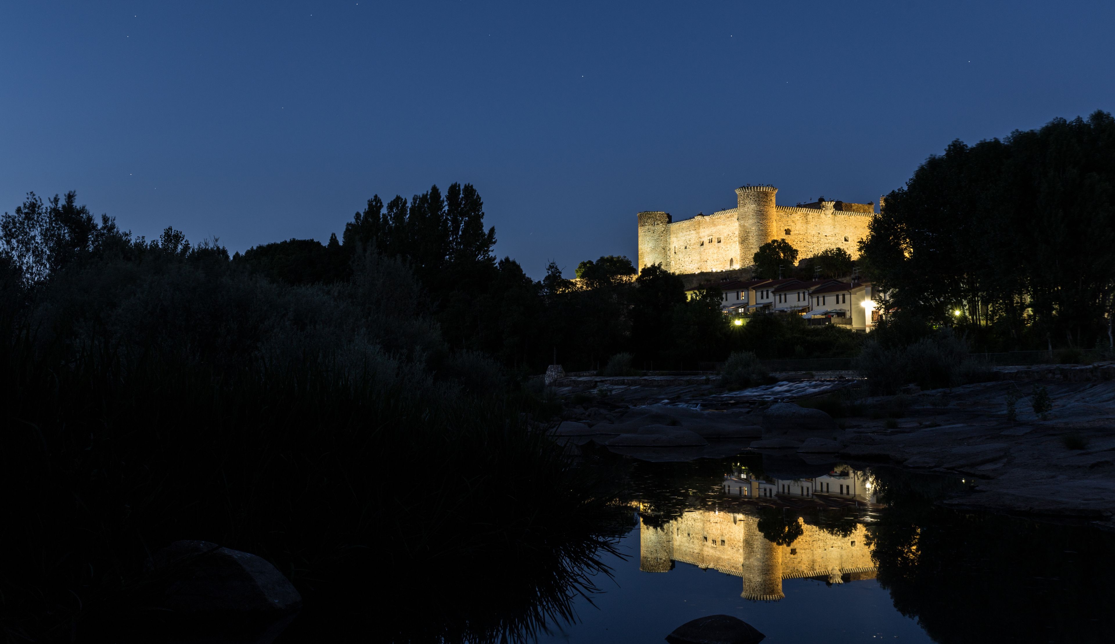 A castle with a moat, illuminated by lights, surrounded by trees and a calm body of water reflecting the structure.