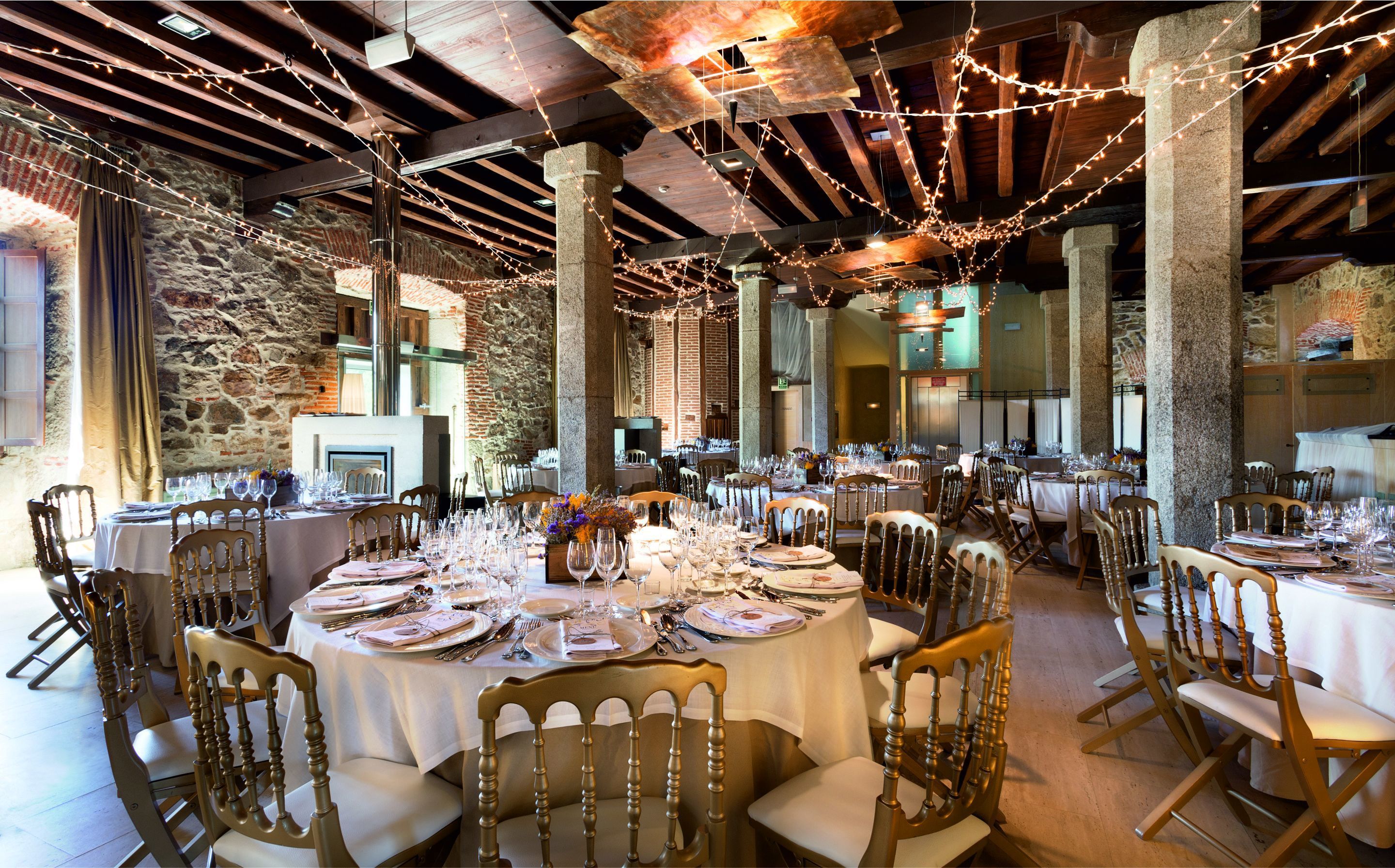 Un salón de banquetes con mesas redondas dispuestas para un evento formal, con paredes de piedra y vigas de madera.