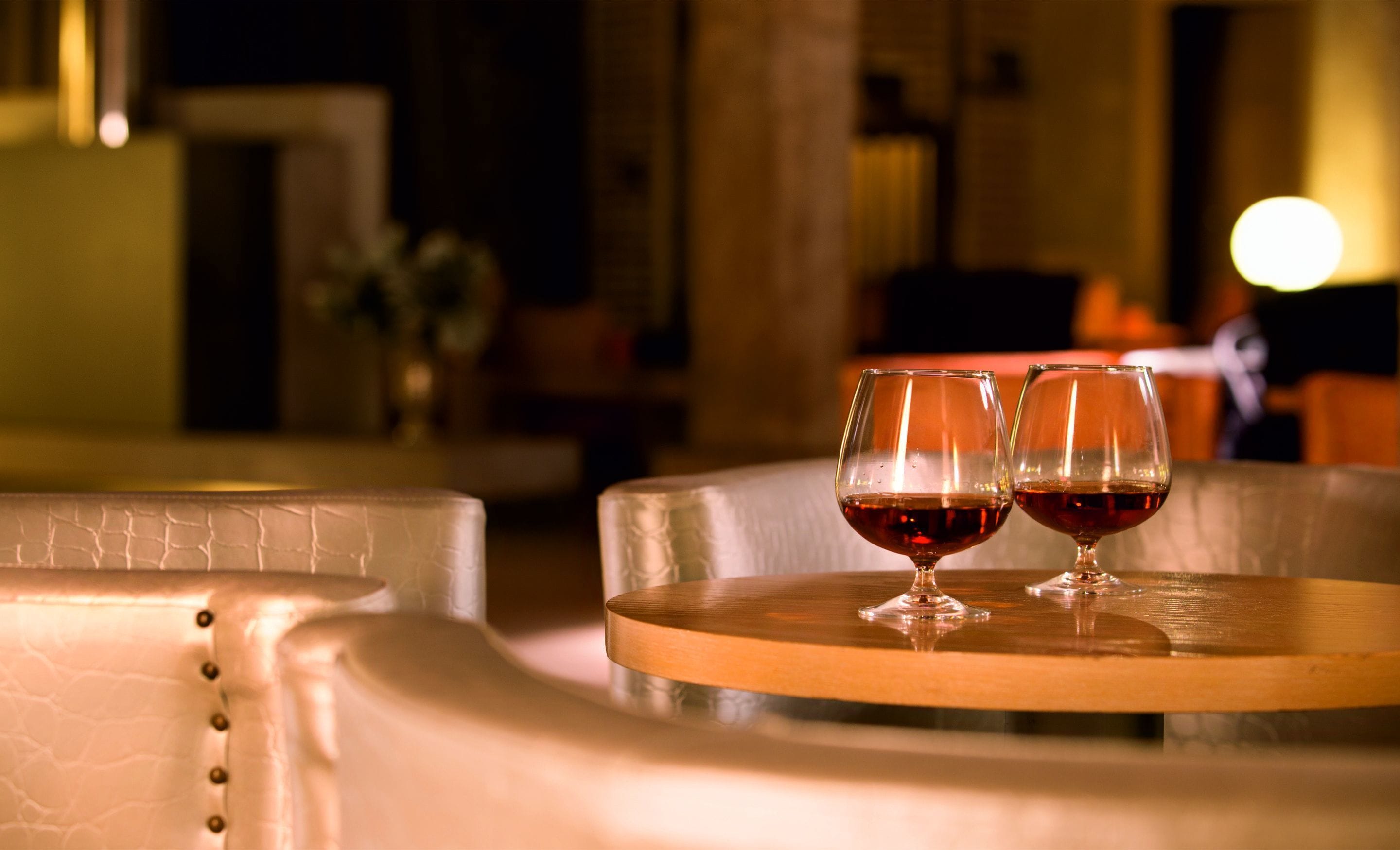 Two wine glasses on a round table with a cushioned chair in a dimly lit room.