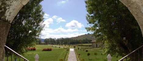 A stone archway leading to a well-maintained garden with a clear pathway, stone balustrades, and a distant view of a building and hills.