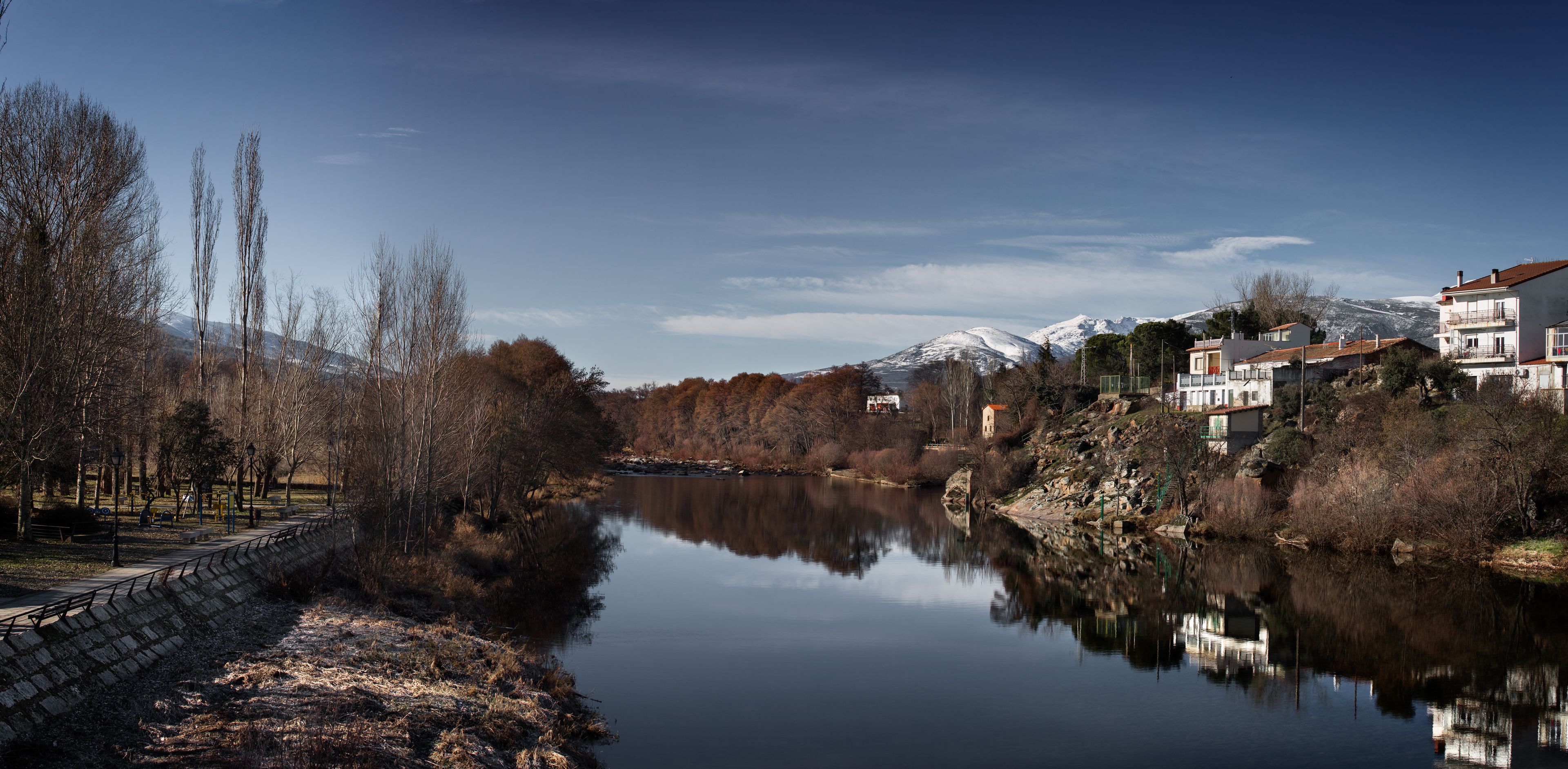 A serene riverside view with a stone wall, trees, and buildings with snow-capped mountains in the background.