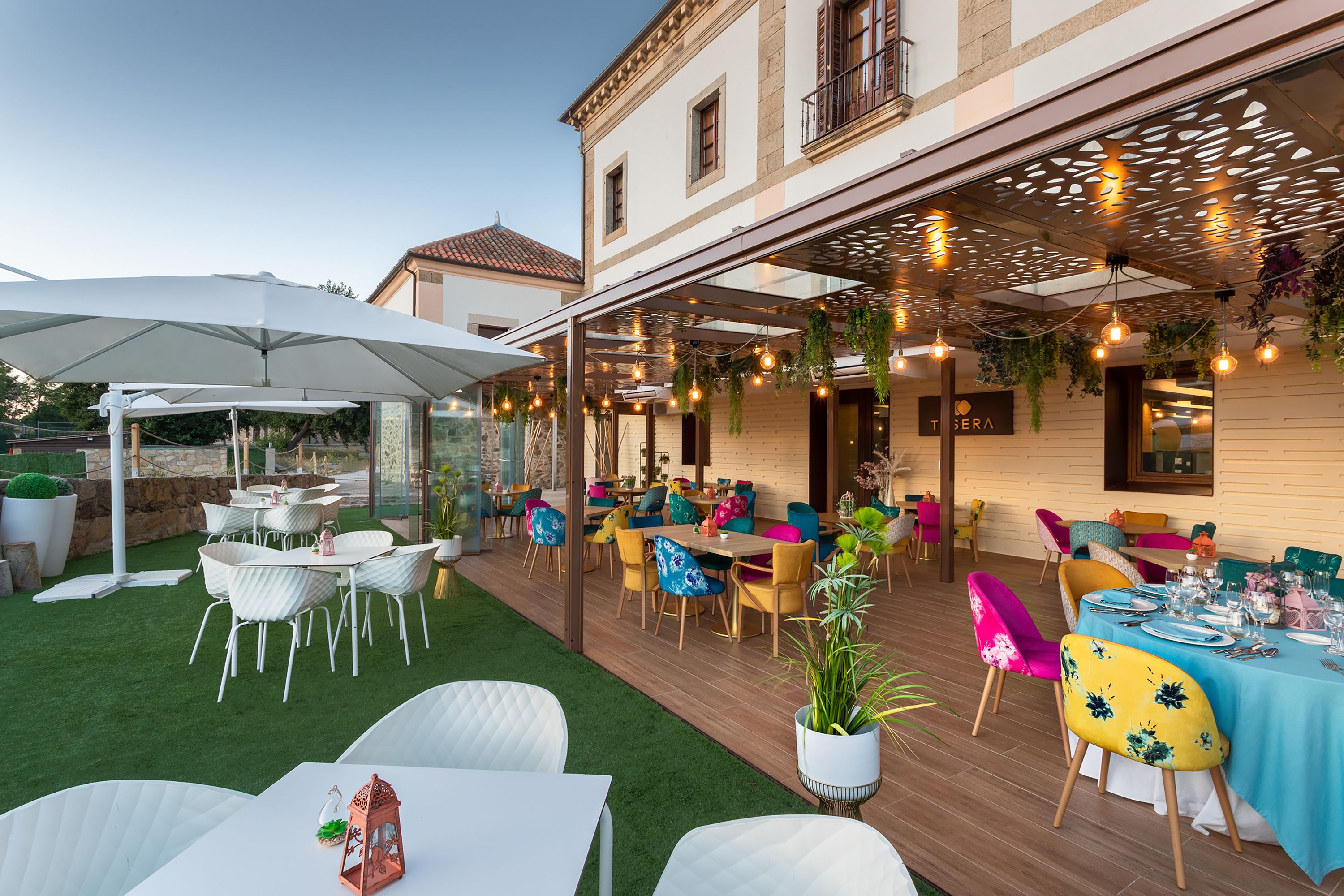Un área de comedor al aire libre con mesas y sillas, una pérgola con luces colgantes y un edificio con balcón al fondo.