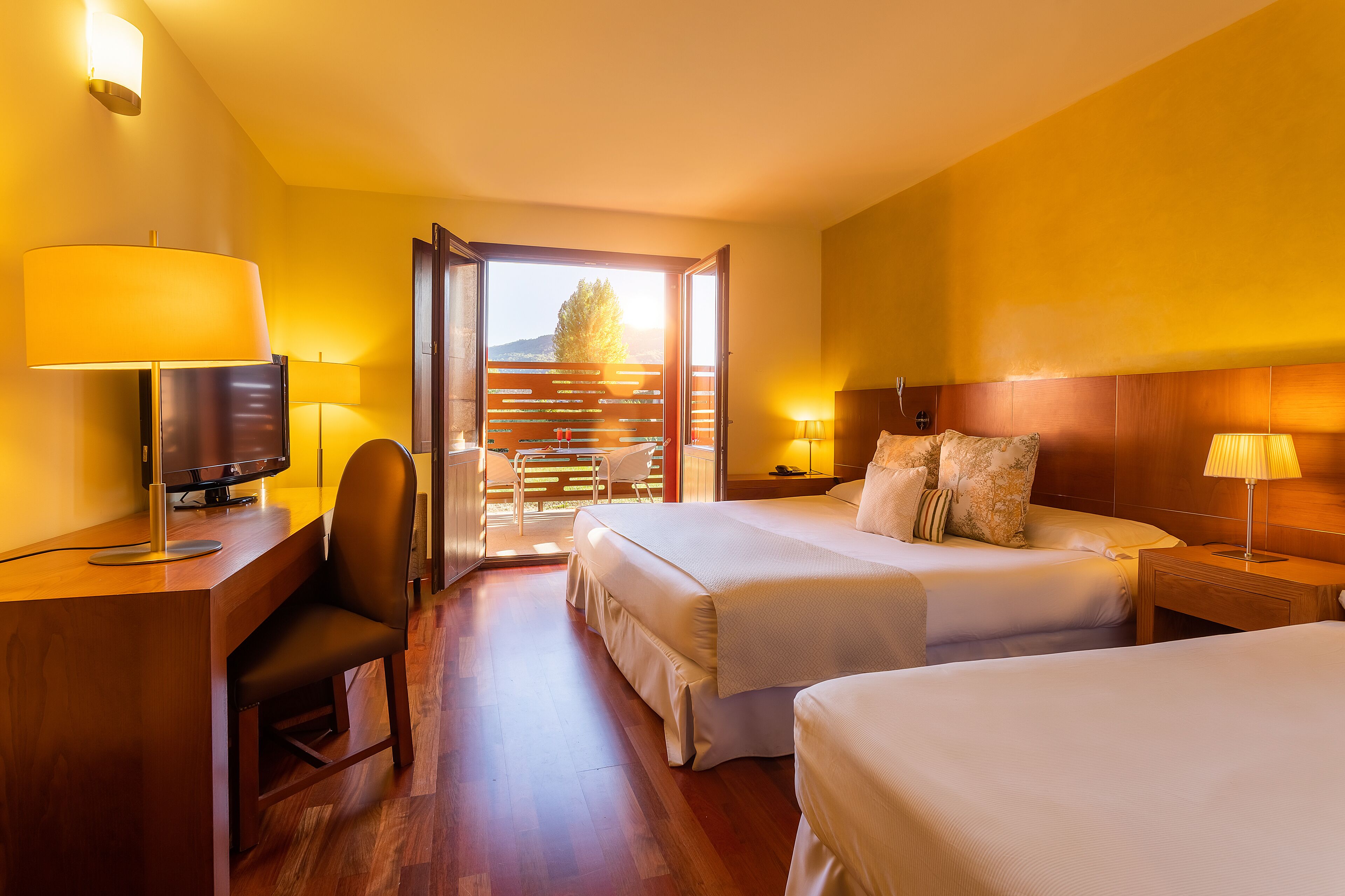 A hotel room with two beds, a desk with a computer, a chair, and a balcony with a view.