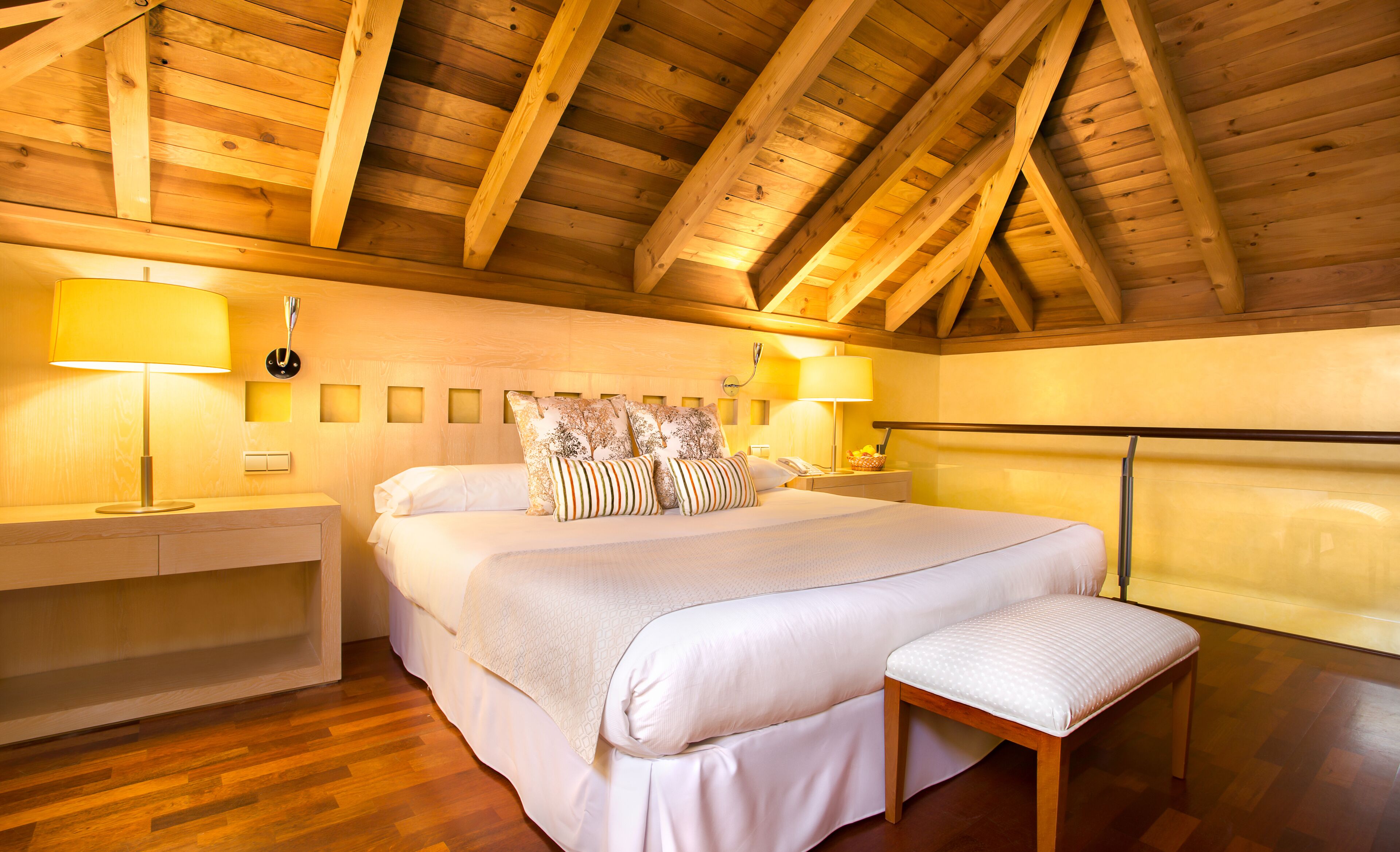 Een houten plafond met schuin ontwerp, een bed met wit beddengoed en gekleurde kussens, twee nachtkastjes met lampen, en een bank aan het voeteneind van het bed.