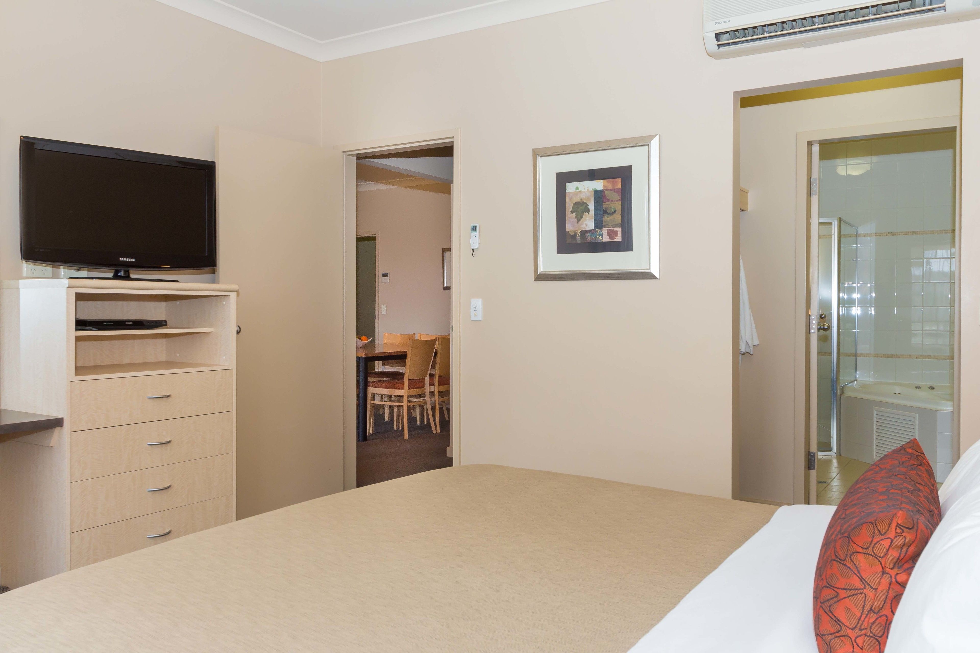 Une chambre d’hôtel avec un lit, un téléviseur posé sur un meuble, un tableau encadré accroché au mur et une salle de bain visible à travers une porte ouverte.