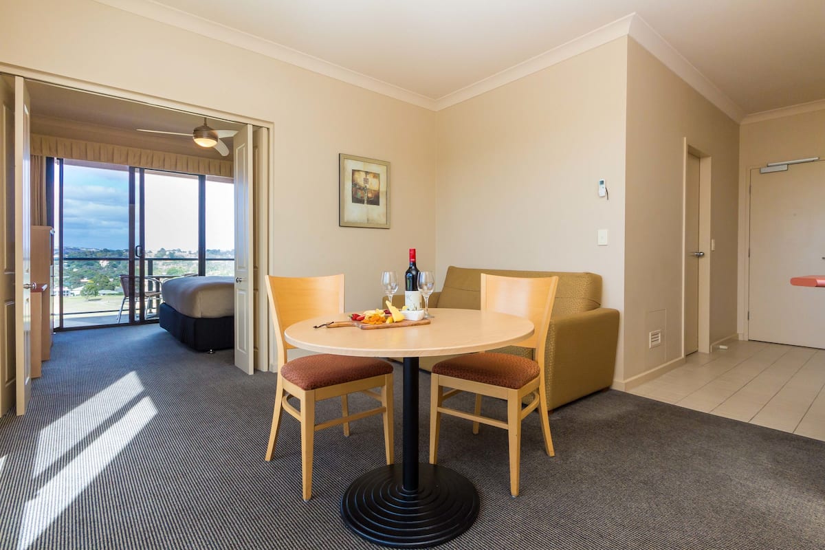 A hotel room with a round table, two chairs, a sofa, a balcony with a view, and a door.