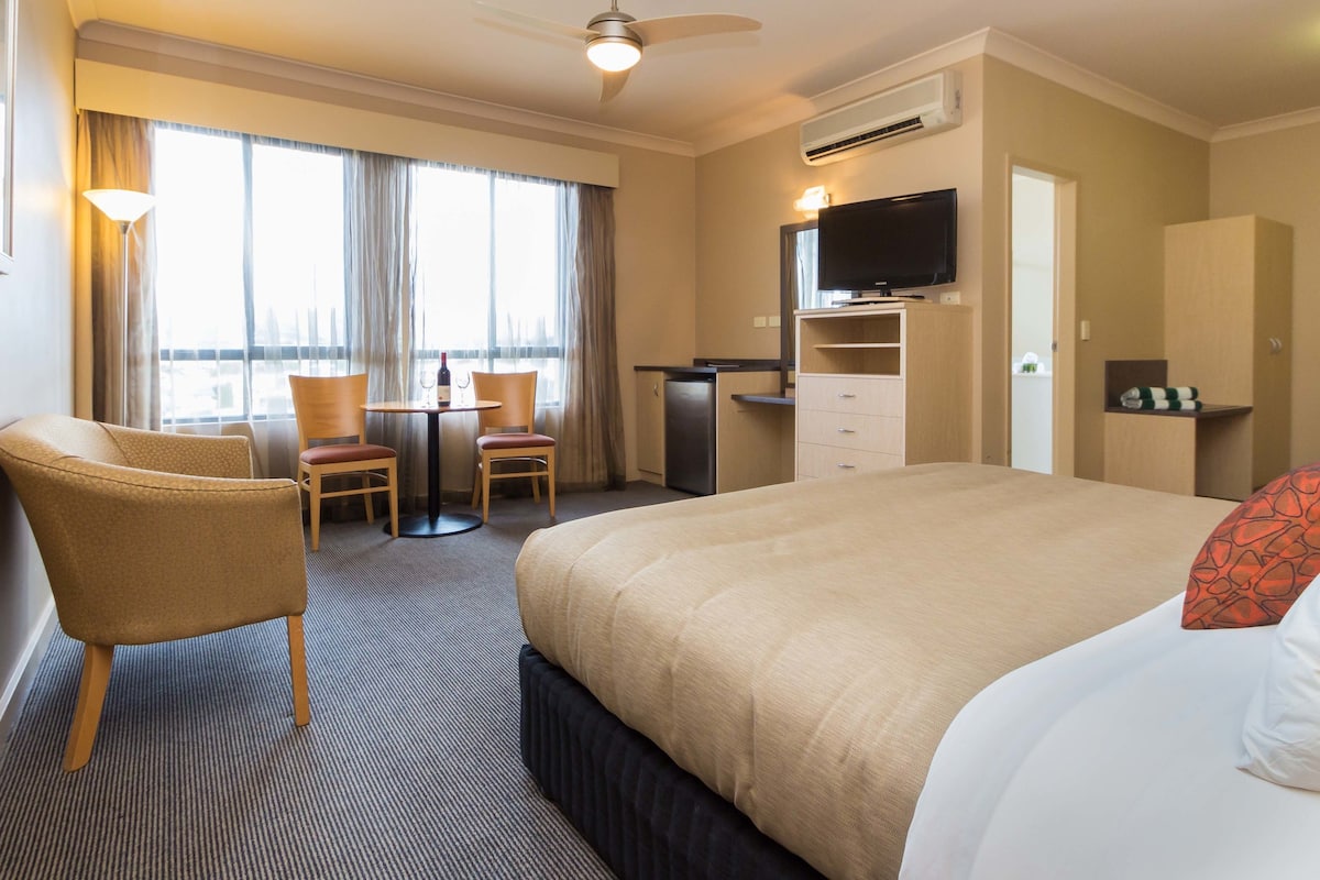 A hotel room with a bed, two chairs, a desk, a television, and an air conditioning unit.