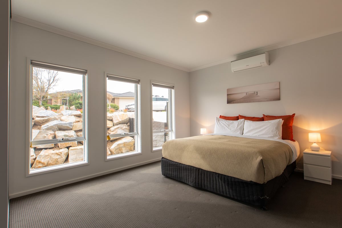 A bedroom with a large bed, three windows, a nightstand, and a wall-mounted air conditioner.