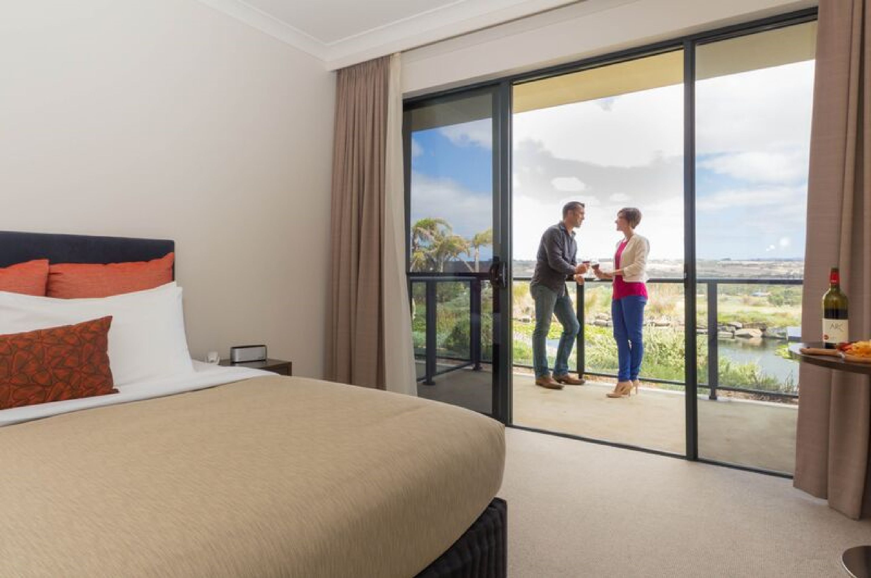 Habitación de hotel con una cama grande, un balcón con vistas a un lago y una mesa con una botella y aperitivos.