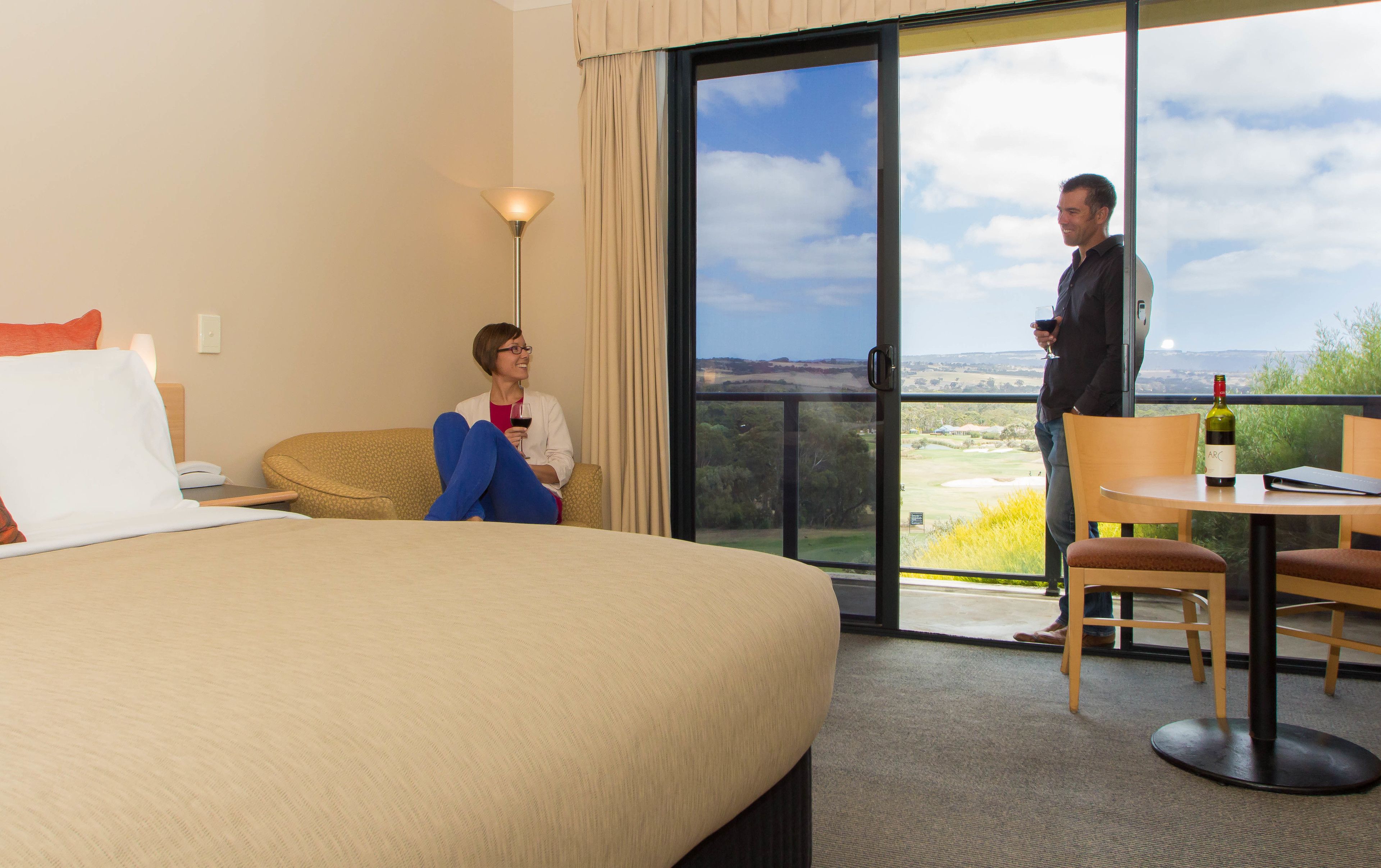 A hotel room with a bed, a sitting area with a lamp, and a balcony with a view of the countryside.