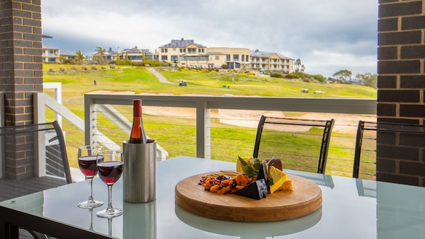Una mesa con una botella de vino, dos copas y una bandeja de frutas en un balcón con vistas a un campo de golf y edificios.