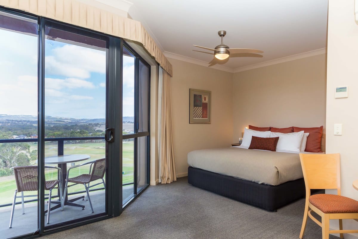 A hotel room with a bed, a ceiling fan, a large window with a view, a small table, and a chair.