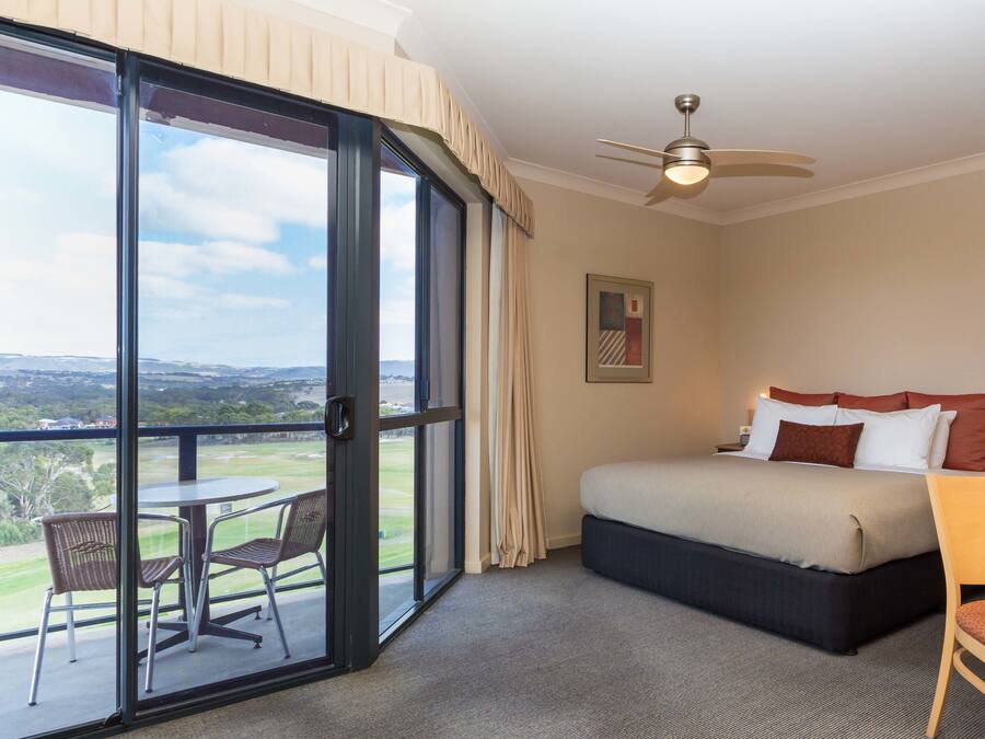 Een hotelkamer met een bed, een plafondventilator, een groot raam met uitzicht, een klein tafeltje en een stoel.