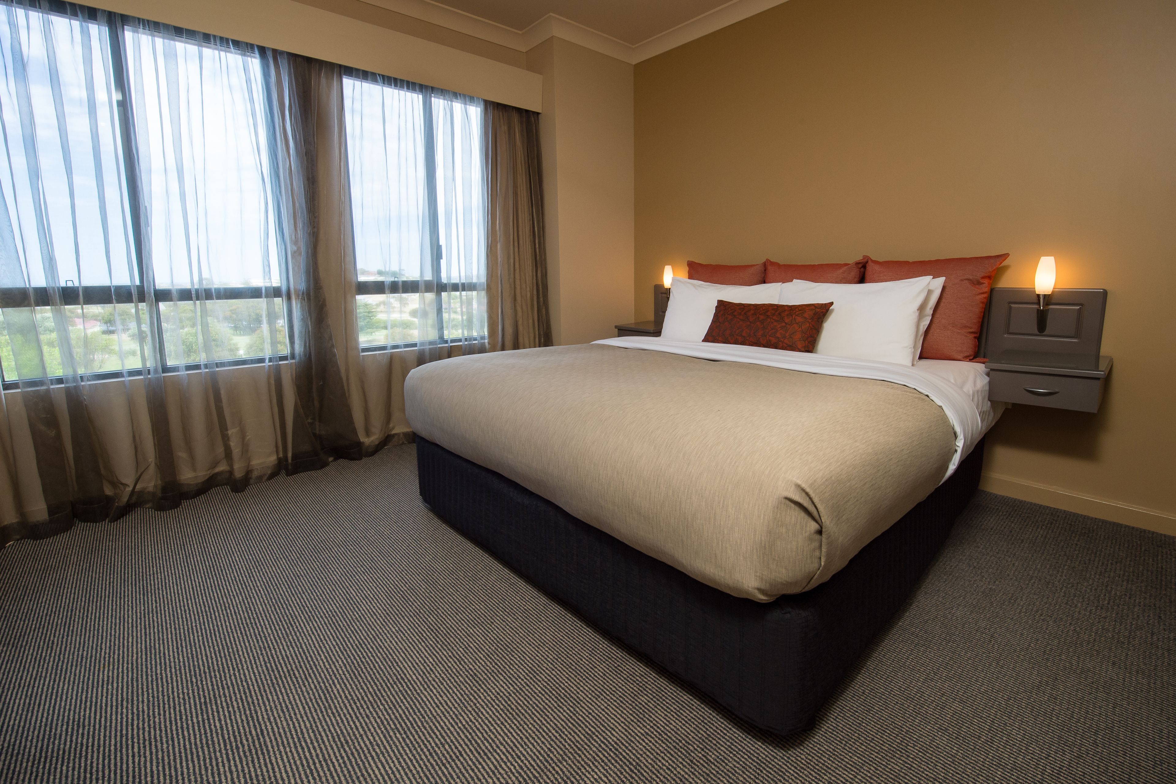 A neatly made bed with a dark headboard, a nightstand with a lamp, and a view of a green landscape through large windows with sheer curtains.