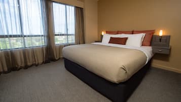 A neatly made bed with a dark headboard, a nightstand with a lamp, and a view of a green landscape through large windows with sheer curtains.