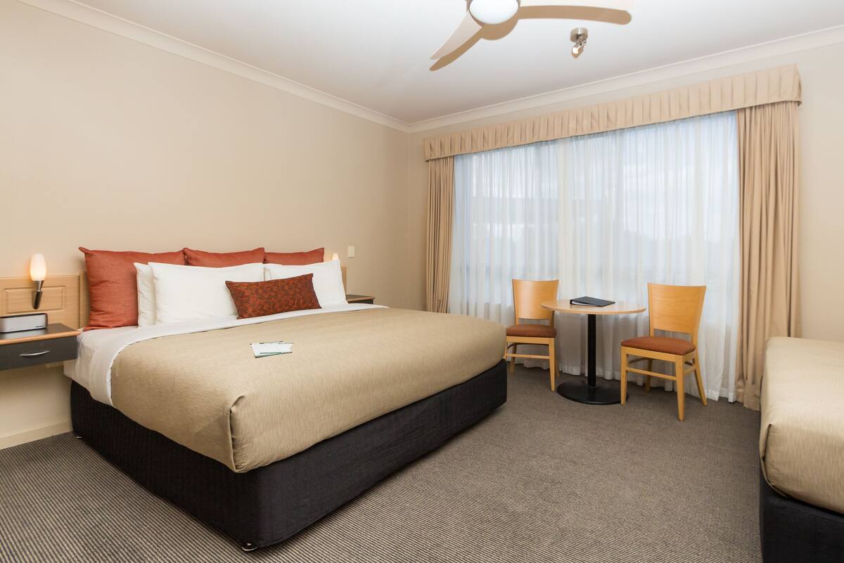 A hotel room with a large bed, a desk, two chairs, a ceiling fan, and a window with curtains.