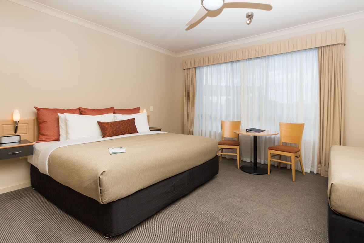 A hotel room with a bed, a desk, two chairs, a ceiling fan, and a large window with curtains.