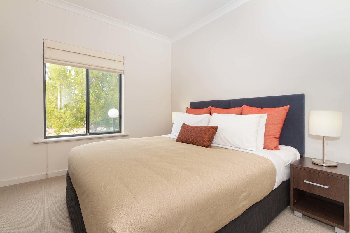 A neatly made bed with a beige blanket and orange pillows, a bedside table with a lamp, and a window with a view of trees.