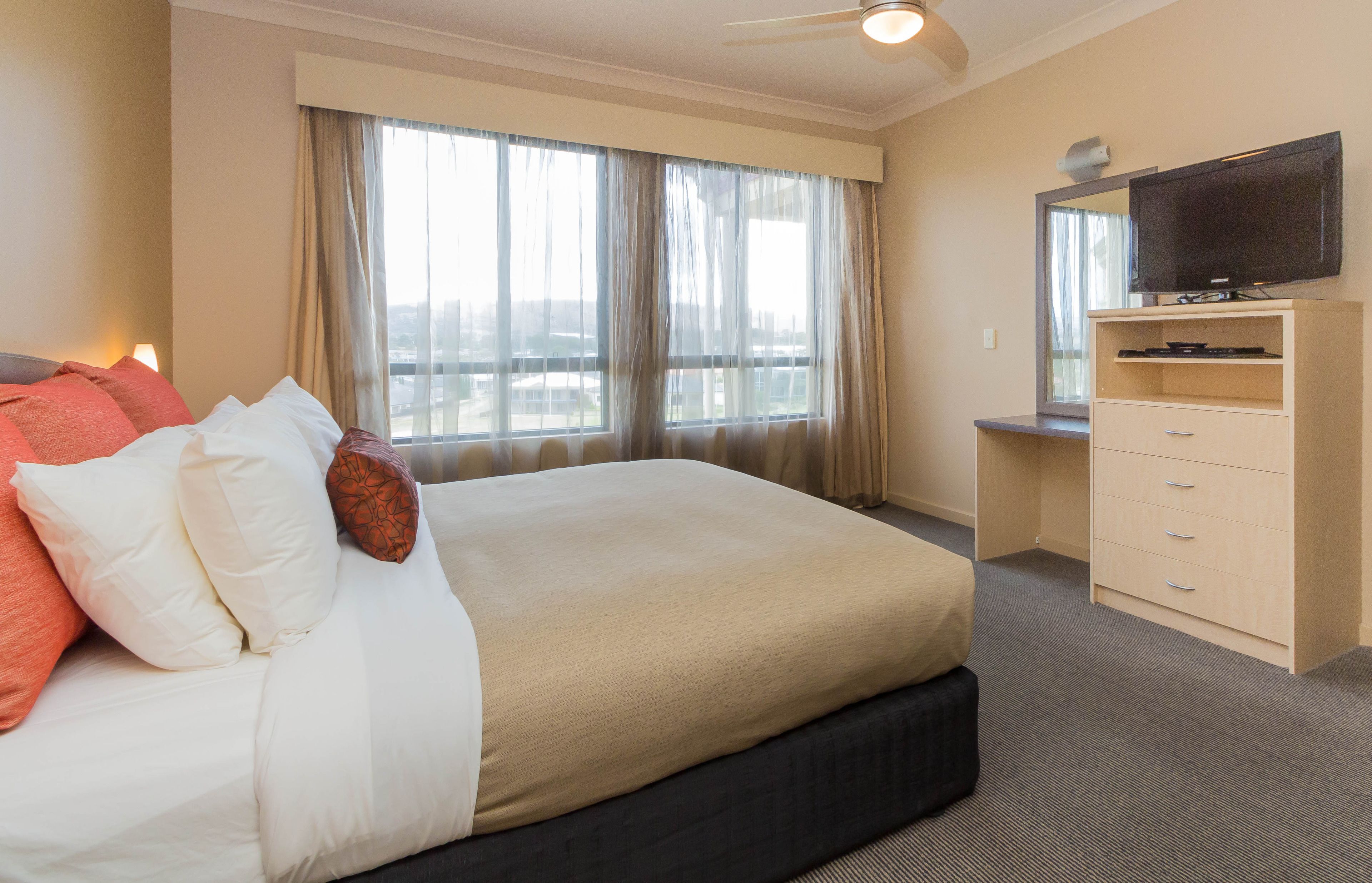 Ein Hotelzimmer mit einem großen Bett, einem Schreibtisch mit Fernseher und einem Vorhangfenster.