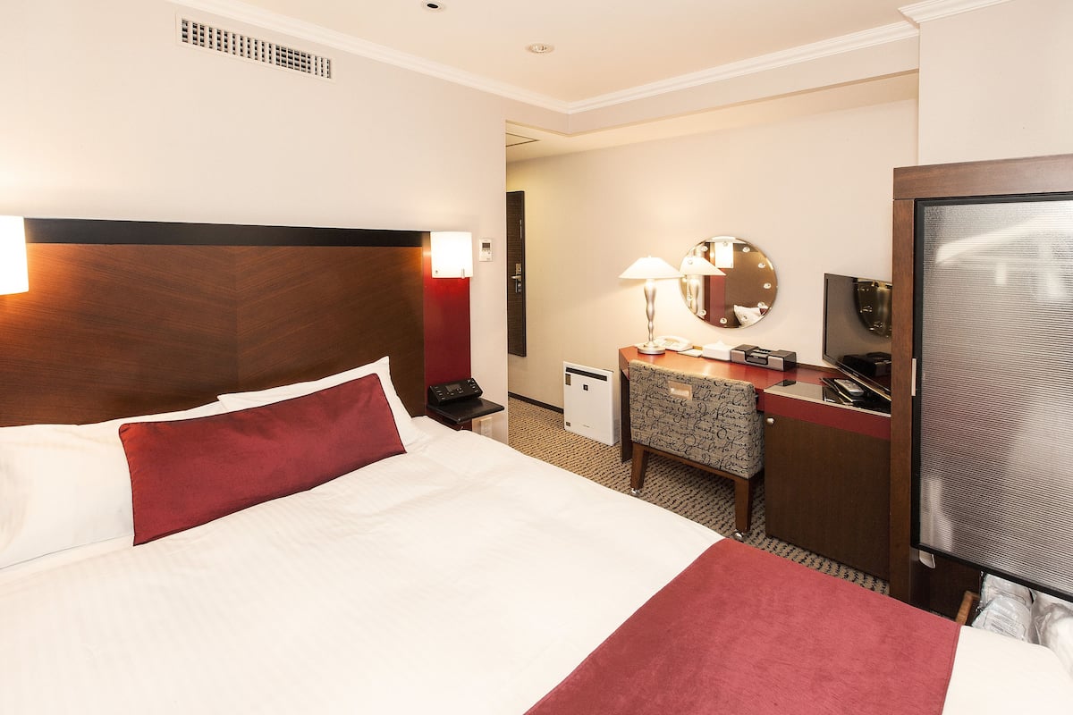 A hotel room with a large bed, a desk with a chair, a mirror, a television, and a small heater.