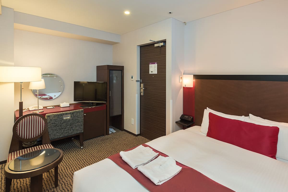 A hotel room with a bed, a desk with a television, a chair, a desk lamp, and a nightstand with a lamp.