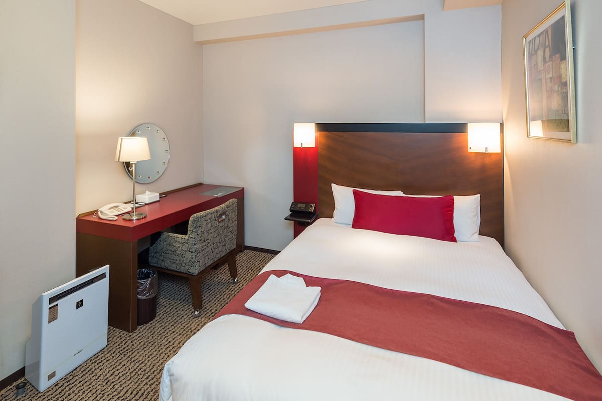 A hotel room with a bed, a desk, a chair, a small table, a telephone, a lamp, and a wall-mounted picture.
