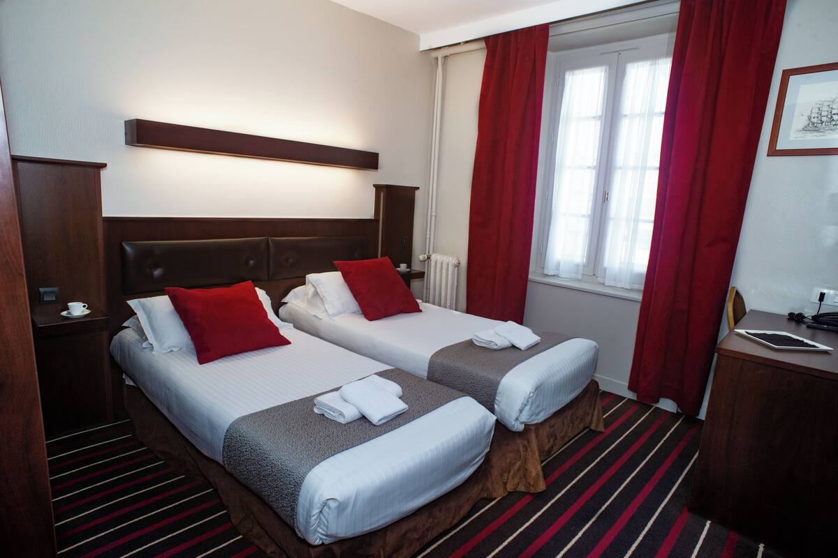 A hotel room with two beds, a nightstand, a desk, and a window with red curtains.