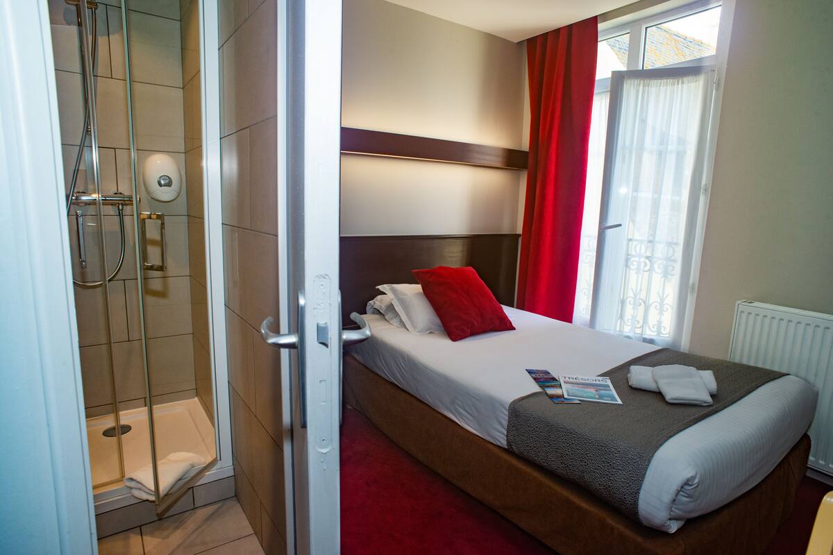 A hotel room with a shower, a bed with a red pillow, a magazine, and a towel on the bed.