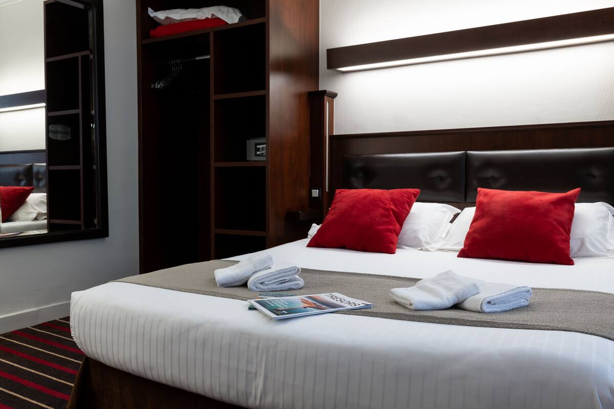 A neatly made bed with red pillows, a magazine, and folded towels.