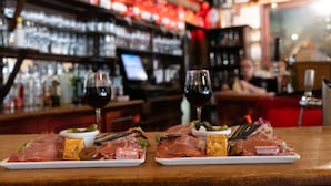 A bar setting with two plates of cured meats and cheese, two glasses of red wine, and a bar counter with bottles and glasses.