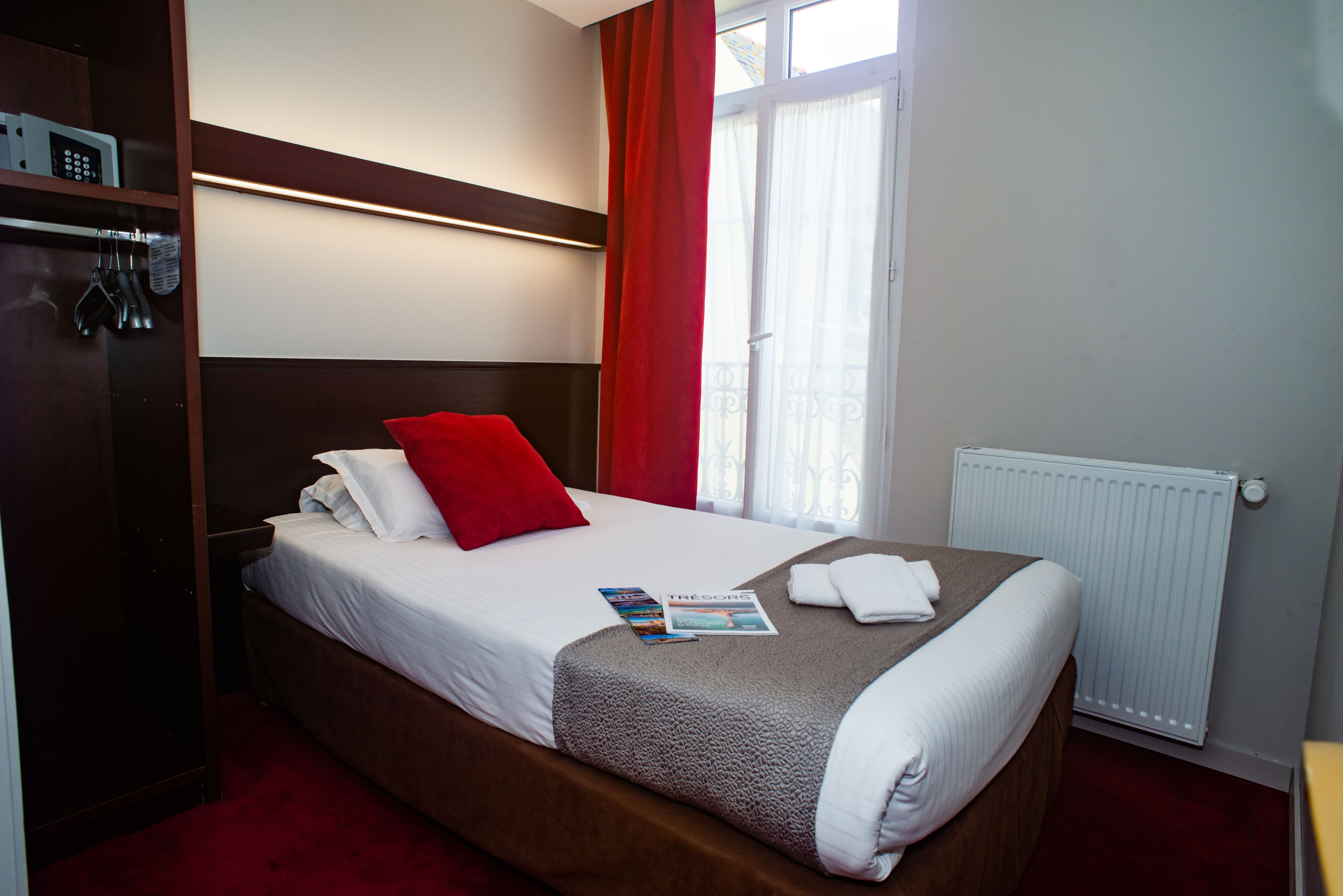 A hotel room with a bed, a red pillow, a magazine, and a white radiator.